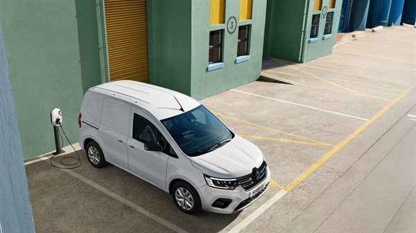 Pogled odozgo na bijelo dostavno vozilo Renault parkirano uz zgradu zelene fasade, spojeno na zidnu punionicu, na označenom parkirnom mjestu.