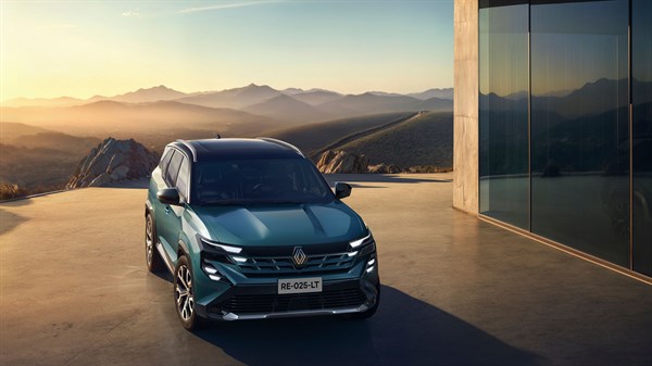Prednji prikaz tamnozelenog vozila Renault parkiranog na terasi uz modernu zgradu sa staklenom fasadom, s planinskim krajolikom u pozadini pri zalasku sunca.