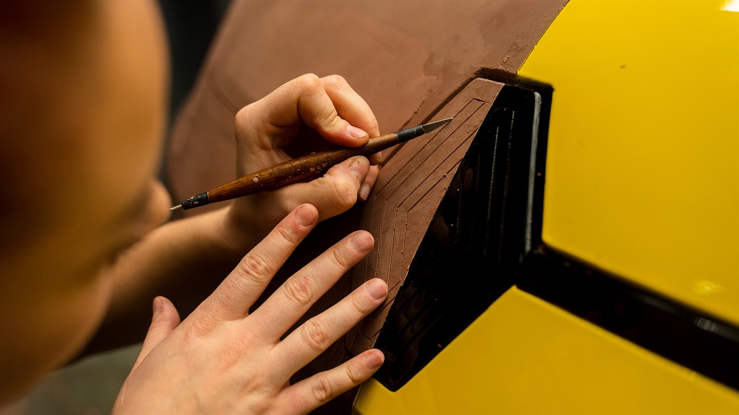 Makro prikaz ruku koje drže alat i oblikuju tamni materijal s vidljivim slojevima i utorima, na žutoj podlozi.