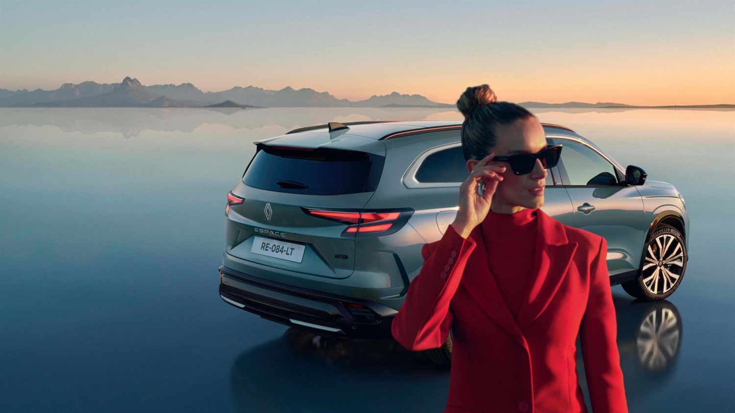 Osoba u crvenom odijelu i sunčanim naočalama stoji ispred vozila Espace full hybrid E-Tech parkiranog na reflektirajućoj površini uz mirno jezero, u pozadini planine i zalazak sunca.