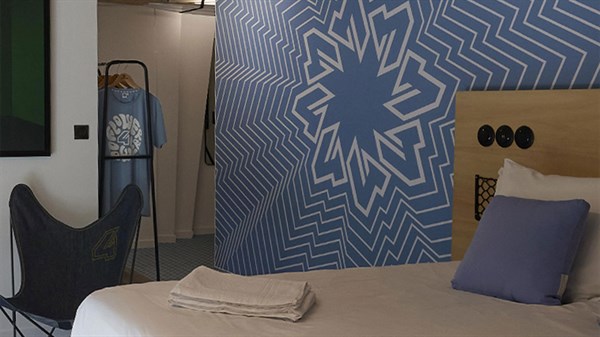 Hotelska soba s plavim zidom s grafičkim uzorkom inspiriranim Renault 4, krevetom i dekorativnim detaljima u modernom stilu.