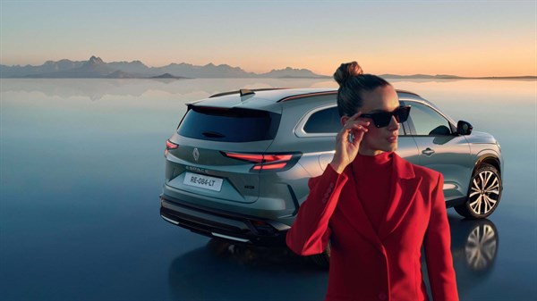 Žena u crvenom odijelu stoji ispred parkiranog vozila Renault uz mirnu vodenu površinu pri zalasku sunca.