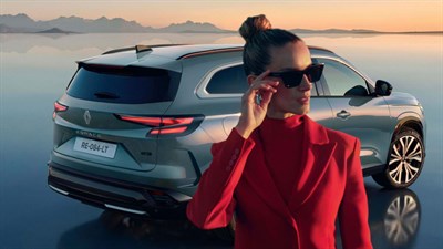 Žena u crvenom odijelu stoji uz vozilo Renault parkirano uz obalu mora u zalazak sunca.
