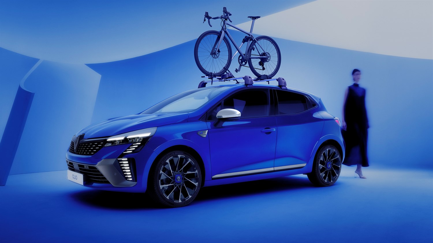 Plavi Clio full hybrid E-Tech s biciklom na krovu parkiran u futurističkom, plavom ambijentu.








