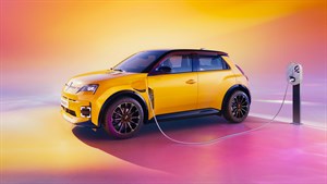 Žuti Renault 5 E-Tech electric spojen na punionicu, prikazan u profilu u živopisnom ambijentu toplih žutih, narančastih i ružičastih tonova.