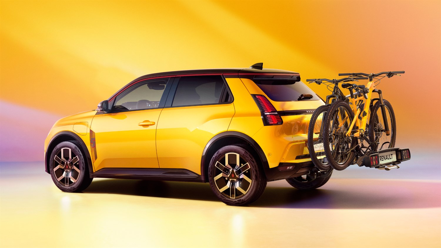 Renault 5 E-Tech electric u žutoj boji prikazan straga s lijeve strane, s montiranim nosačem i dva bicikla, na pozadini u toplim žuto-narančastim tonovima
