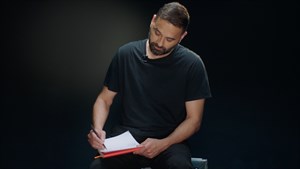 Muškarac sjedi i piše na papiru u studijskom ambijentu, snimka iz R:talk videa

